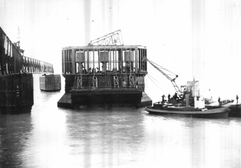 Caisson P.4, échouage au nord de son point d'échouage.
La mer descendue à +1 laisse voir le dessous des flotteurs
et la poutre d'accrochage.
Le ponton-grue embarque des pompes pour déséchouer le caisson
et l'amener en place à marée haute.